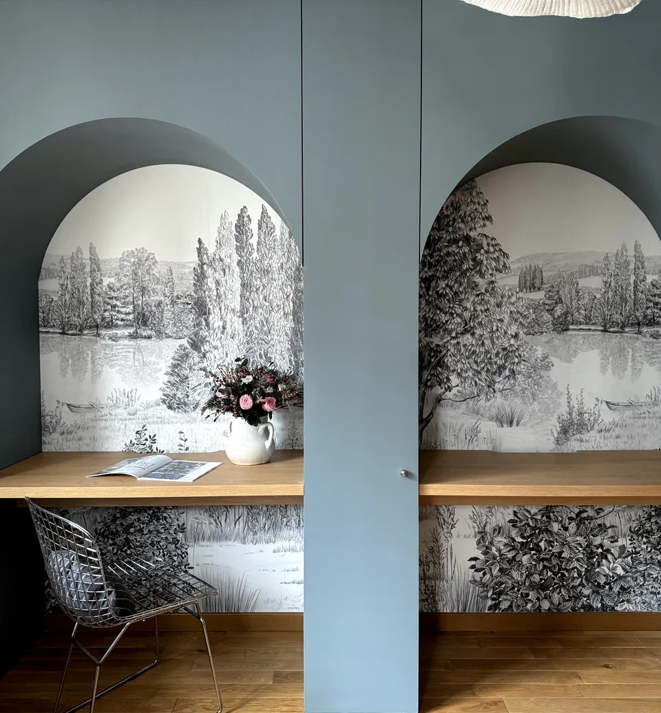 Studio Madame : architecte d’intérieur, Paris et Montmorency
Projet réalisé à Enghien-les-Bains.
Photographie d’intérieur : aménagement sur mesure bleu de Nimes (Farrow & Ball) composé de colonnes et d’arches intégrant deux espaces bureau nichés entre les éléments. Un papier peint sous forme de fresque en noir et blanc tapisse le fond des niches.
Sur l’un des plateaux en chêne du bureau, un magazine ainsi qu’un bouquet de fleurs dans des tons de rose sont posés. Une chaise à quadrillage métallique est placée devant ce bureau.