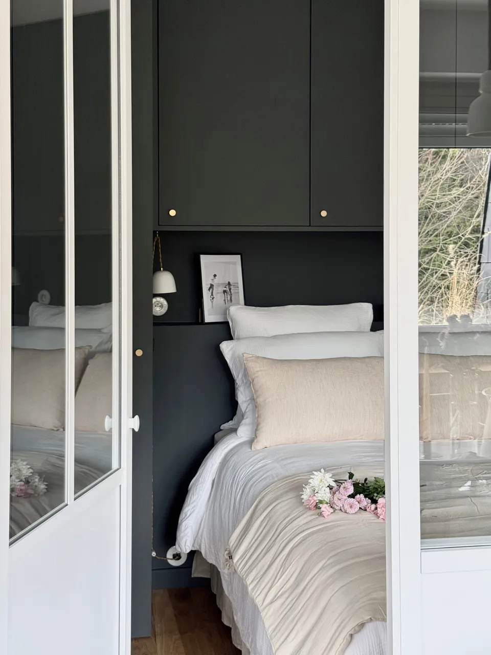 Studio Madame : architecte d’intérieur, Paris et Montmorency
Projet réalisé à Montmorency.
Photographie d’intérieur : chambre parentale avec dressing réalisé sur mesure.
Le dressing est peint dans un gris-vert foncé (Down Pipe - F&B).
Le lit est habillé d’une parure blanche en gaze de coton, de coussins et édredon beiges.
La prise ainsi que les baladeuses sont en porcelaine.
Des fleurs sont posées sur le lit.
La chambre est visible en transparence grâce à une verrière atelier.