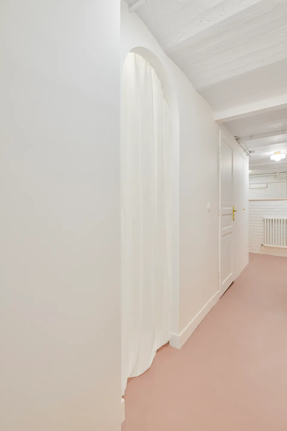 Studio Madame : architecte d’intérieur, Paris et Montmorency.
Projet réalisé à Deuil-la-Barre.
Photographie d’intérieur : couloir entièrement repeint en blanc (Ressource) amenant sur une petite salle de bain par un passage en arche habillé d’un rideau blanc. Le sous-sol présente toutes les caractéristiques typiques d’une meulière (plafond voûté, tuyaux apparents, briques, etc.). Les murs sont peints en blanc (Ressource) et le sol est habillé d’un béton ciré rose (Harmony Béton).