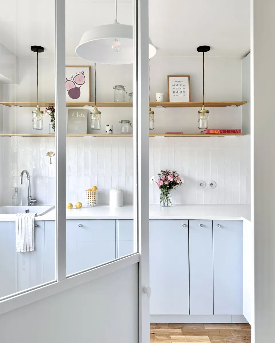 Studio Madame : architecte d’intérieur, Paris et Montmorency
Projet réalisé à Montmorency.
Photographie d’intérieur : cuisine composée de façades bleu ciel et d’un plan de travail blanc.
La cuisine est habillée d’une faïence artisanale blanche et d’un parquet en chêne.
Quatre suspensions bocal tombent au-dessus du plan de travail, à la hauteur d’étagères en chêne sur lesquelles reposent divers objets de décoration (livres, cadres, fleurs).
La cuisine est visible en transparence grâce à une verrière atelier.