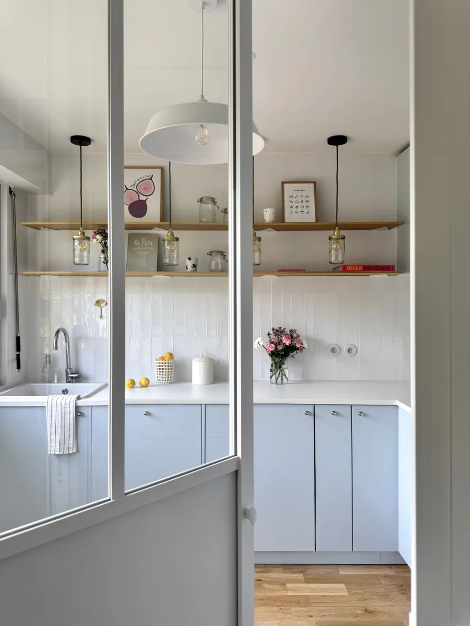 Studio Madame : architecte d’intérieur, Paris et Montmorency
Projet réalisé à Montmorency.
Photographie d’intérieur : cuisine composée de façades bleu ciel et d’un plan de travail blanc.
La cuisine est habillée d’une faïence artisanale blanche et d’un parquet en chêne.
Quatre suspensions bocal tombent au-dessus du plan de travail, à la hauteur d’étagères en chêne sur lesquelles reposent divers objets de décoration (livres, cadres, fleurs).
La cuisine est visible en transparence grâce à une verrière atelier.