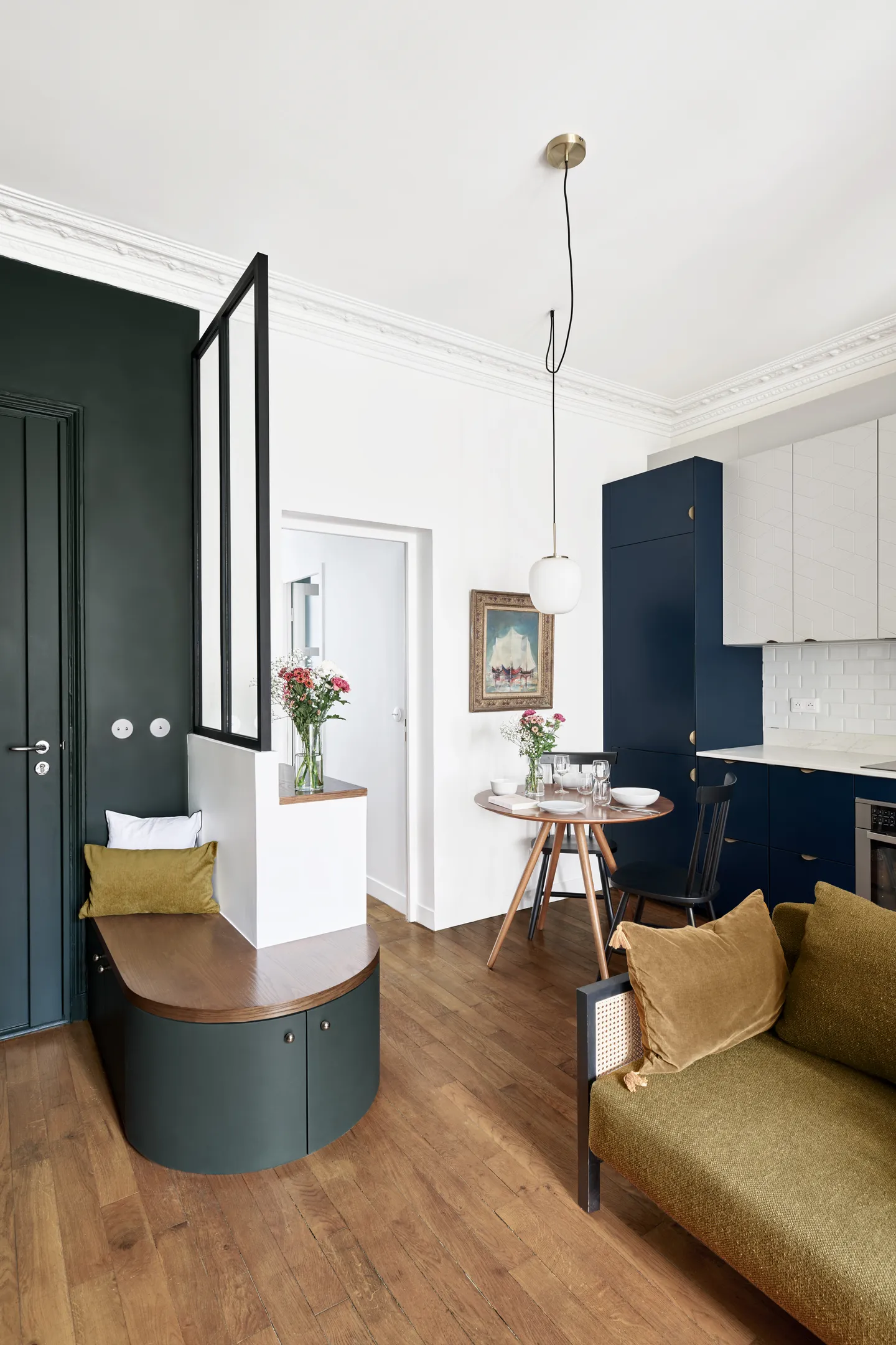 Studio Madame : architecte d’intérieur, Paris et Montmorency.
Projet réalisé à Boulogne-Billancourt. 
Photographie d’intérieur : entrée entièrement peinte en vert foncé (Ressource) et donnant sur un espace ouvert avec salon, salle à manger et cuisine.
L’entrée est composée d’une banquette arrondie réalisée sur mesure et d’une verrière noire. Le reste de l’espace comprend un canapé couleur bronze, une table à manger noyer avec deux chaises noires et une cuisine blanche et bleu nuit.
Les autres murs sont peints en blanc (Ressource) et un parquet massif ancien est au sol.