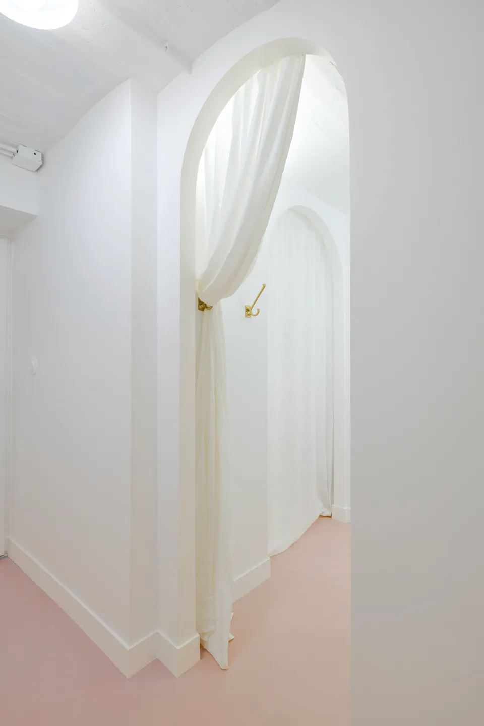 Studio Madame : architecte d’intérieur, Paris et Montmorency.
Projet réalisé à Deuil-la-Barre.
Photographie d’intérieur : couloir entièrement repeint en blanc (Ressource) amenant sur une petite salle de bain par un passage en arche habillé d’un rideau blanc. Le sous-sol présente toutes les caractéristiques typiques d’une meulière (plafond voûté, tuyaux apparents, briques, etc.). Les murs sont peints en blanc (Ressource) et le sol est habillé d’un béton ciré rose (Harmony Béton).