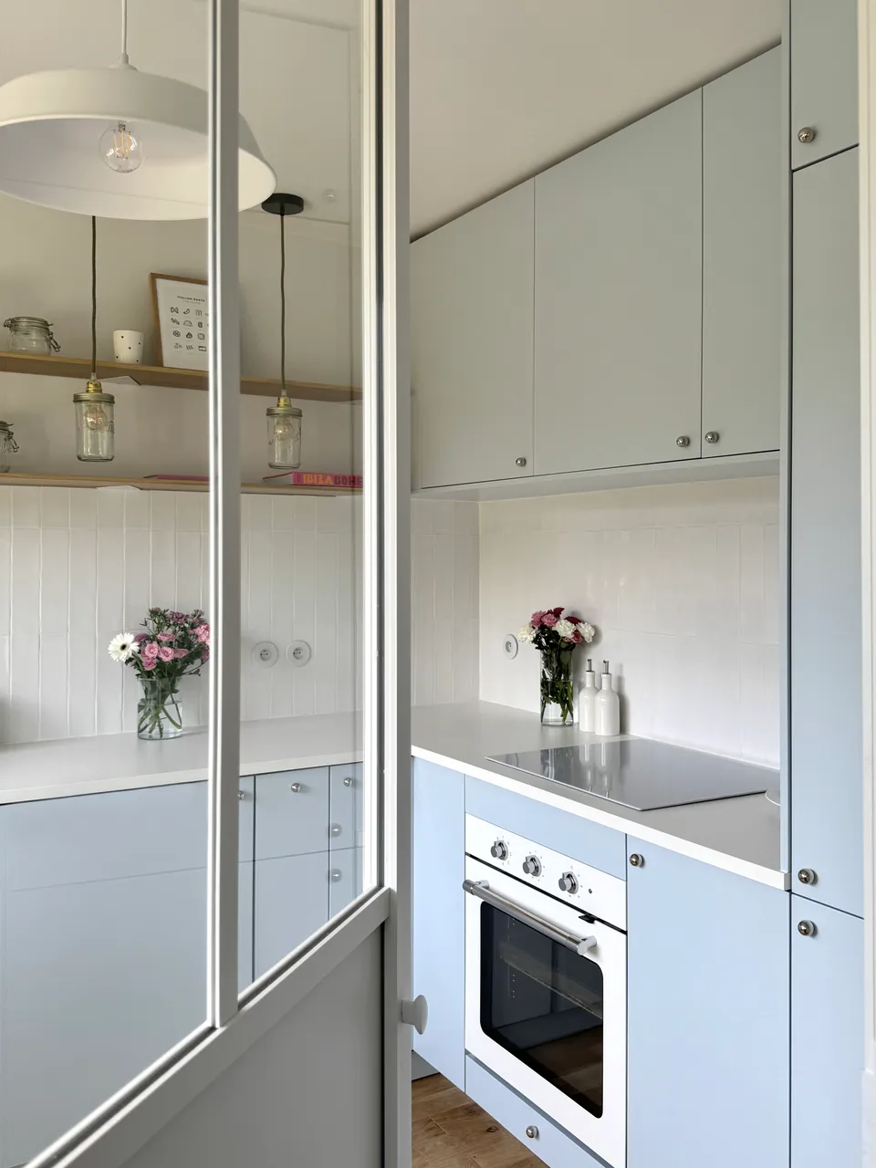 Studio Madame : architecte d’intérieur, Paris et Montmorency
Projet réalisé à Montmorency.
Photographie d’intérieur : cuisine composée de façades bleu ciel et d’un plan de travail blanc.
La cuisine est habillée d’une faïence artisanale blanche et d’un parquet en chêne.
Quatre suspensions bocal tombent au-dessus du plan de travail, à la hauteur d’étagères en chêne sur lesquelles reposent divers objets de décoration (livres, cadres, fleurs).
La cuisine est visible en transparence grâce à une verrière atelier.