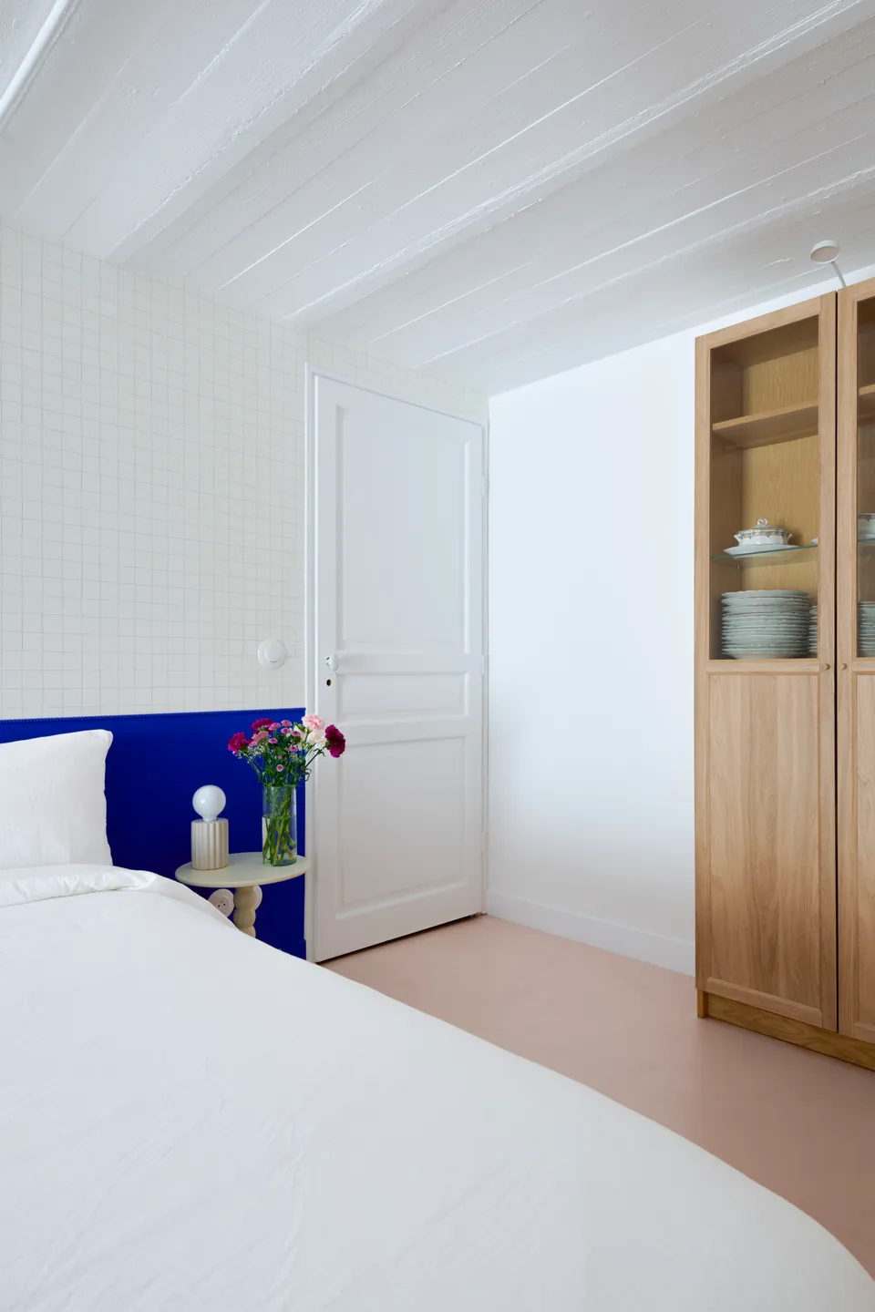 Studio Madame : architecte d’intérieur, Paris et Montmorency.
Projet réalisé à Deuil-la-Barre.
Photographie d’intérieur : soubassement (tête de lit) peint en bleu Klein (Ressource) intégrant un espace nuit agrémenté d’un papier peint minimaliste quadrillé (Season Paper), d’une lampe et table de chevet beiges aux lignes originales et d’un lit avec parure blanche. Une bibliothèque vitrée en chêne permet d’inclure des rangements supplémentaires. Le plafond voûté est typique des sous-sols présents dans les meulières. Les murs sont peints en blanc et bleu Klein (Ressource) et le sol est habillé d’un béton ciré rose (Harmony Béton).