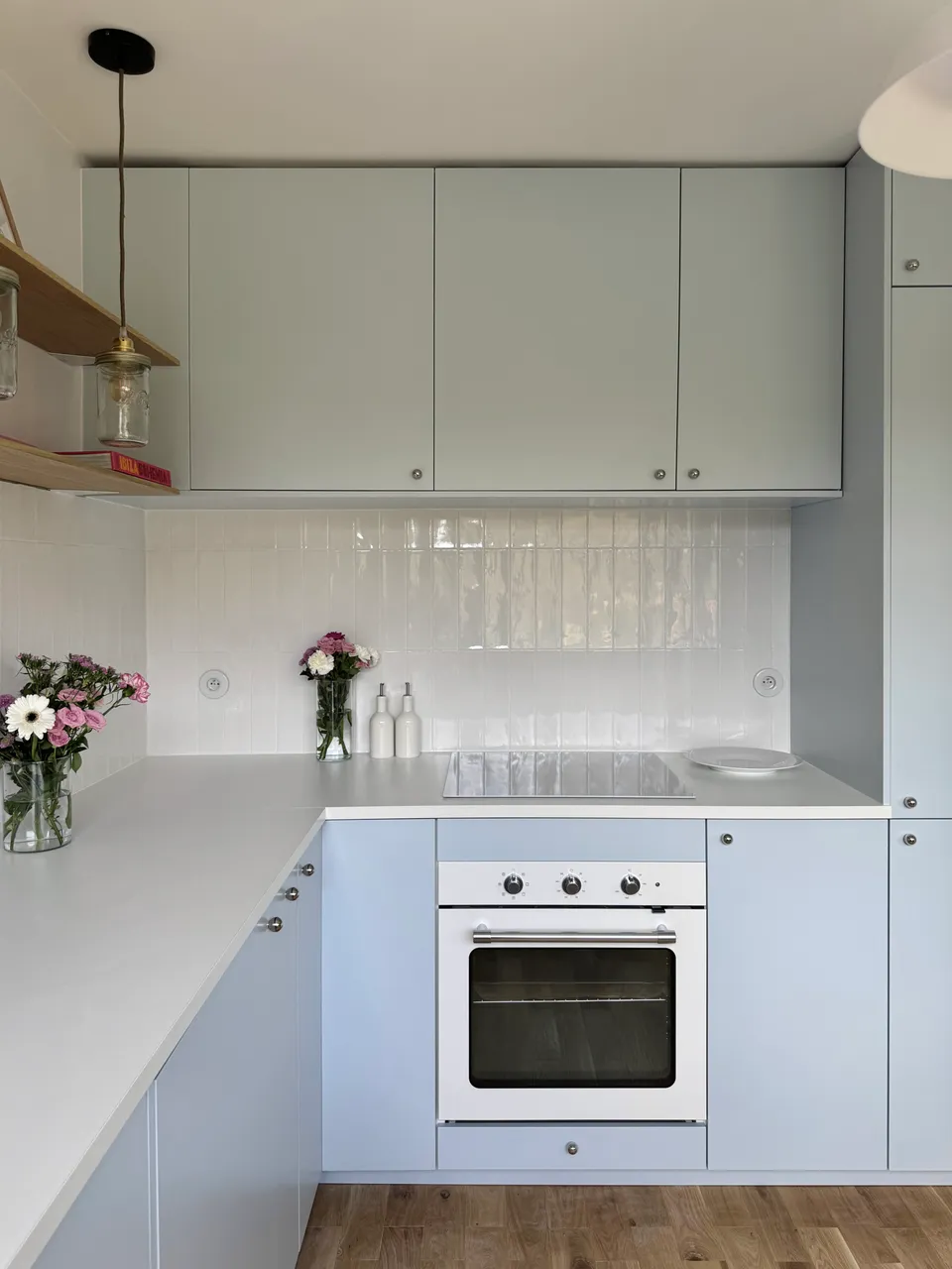 Studio Madame : architecte d’intérieur, Paris et Montmorency
Projet réalisé à Montmorency.
Photographie d’intérieur : cuisine composée de façades bleu ciel et d’un plan de travail blanc.
La cuisine est habillée d’une faïence artisanale blanche et d’un parquet en chêne.
Quatre suspensions bocal tombent au-dessus du plan de travail, à la hauteur d’étagères en chêne sur lesquelles reposent divers objets de décoration (livres, cadres, fleurs).
La cuisine est visible en transparence grâce à une verrière atelier.