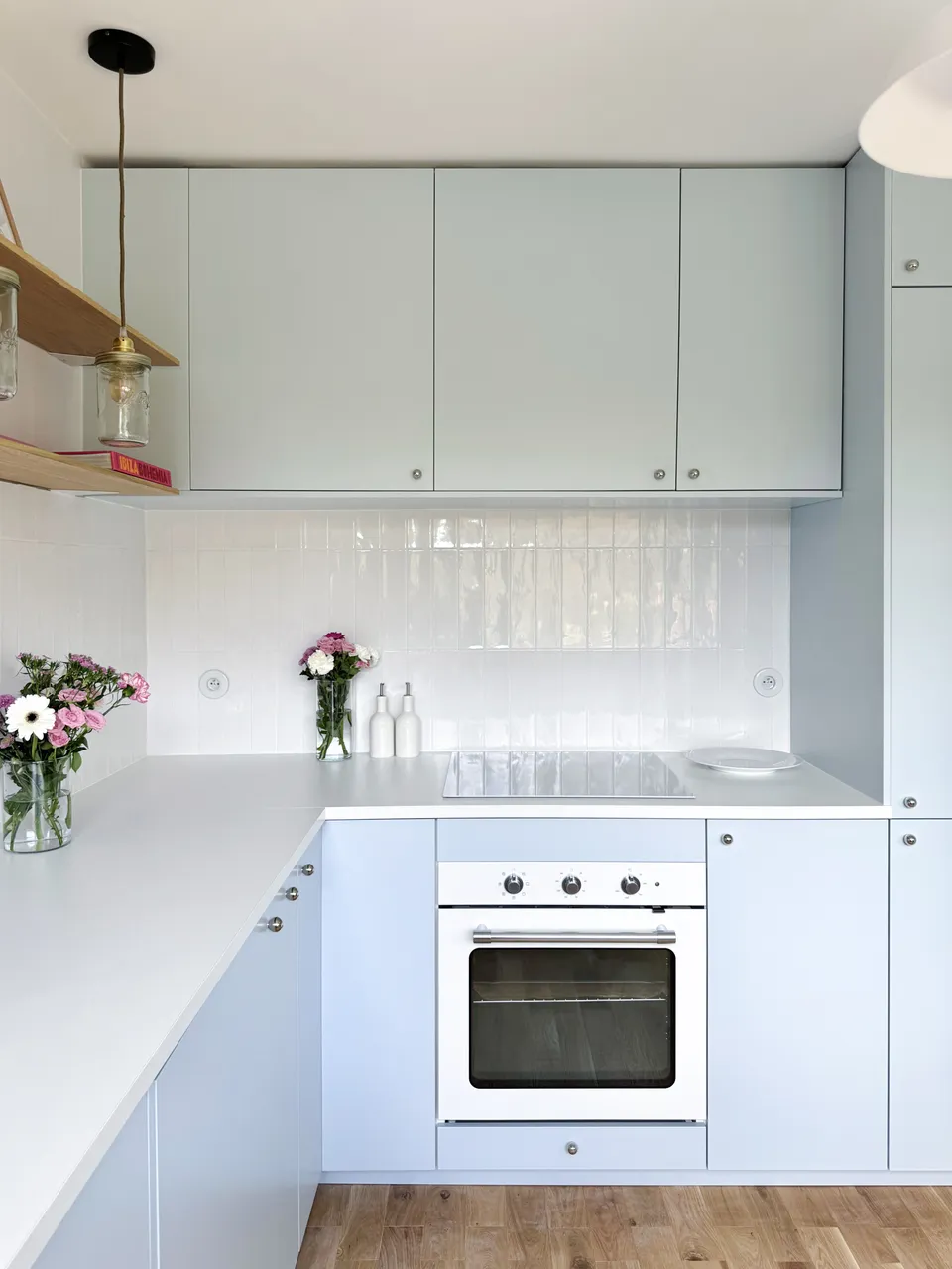 Studio Madame : architecte d’intérieur, Paris et Montmorency
Projet réalisé à Montmorency.
Photographie d’intérieur : cuisine composée de façades bleu ciel et d’un plan de travail blanc.
La cuisine est habillée d’une faïence artisanale blanche et d’un parquet en chêne.
Quatre suspensions bocal tombent au-dessus du plan de travail, à la hauteur d’étagères en chêne sur lesquelles reposent divers objets de décoration (livres, cadres, fleurs).
La cuisine est visible en transparence grâce à une verrière atelier.