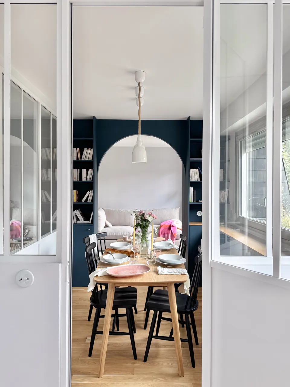 Studio Madame : architecte d’intérieur, Paris et Montmorency
Projet réalisé à Montmorency.
Photographie d’intérieur : salle à manger avec vue sur salon, composée d’une table rectangulaire avec 4 chaises vintages noires. La table est dressée et des suspensions tombent au-dessus de la table. Le salon est visible grâce à la bibliothèque sur mesure et son passage en arche.