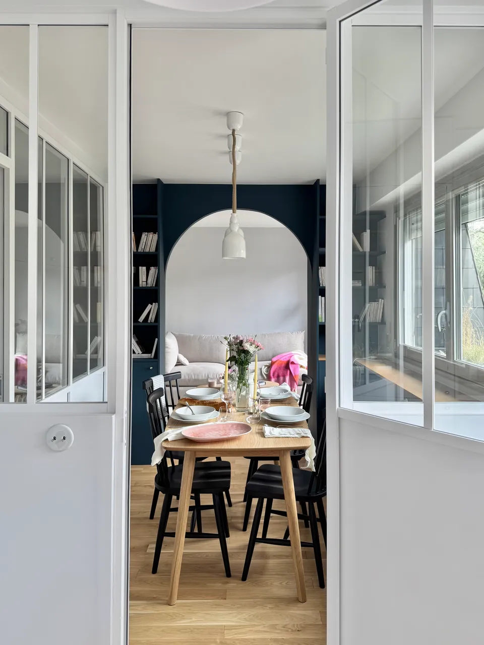Studio Madame : architecte d’intérieur, Paris et Montmorency
Projet réalisé à Montmorency.
Photographie d’intérieur : salle à manger avec vue sur salon, composée d’une table rectangulaire avec 4 chaises vintages noires. La table est dressée et des suspensions tombent au-dessus de la table. Le salon est visible grâce à la bibliothèque sur mesure et son passage en arche.