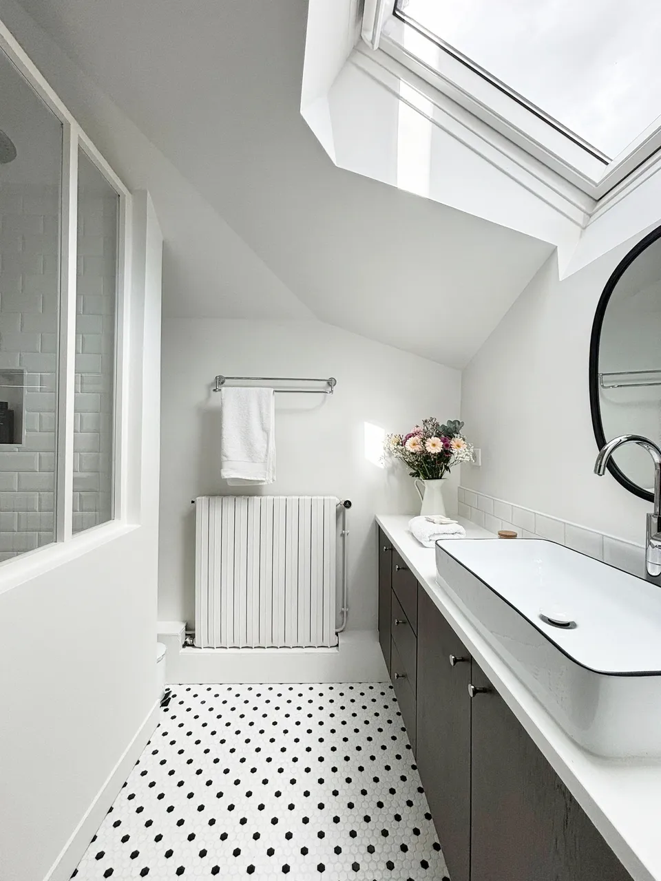 Studio Madame : architecte d’intérieur, Paris et Montmorency
Projet réalisé à Cormeilles-en-Parisis.
Photographie d’intérieur : salle de bain avec douche séparée par une verrière blanche, carreaux de métro en faïence, sol en mosaïque hexagonale blanche à pois noirs, meuble vasque en noyer et vasque en céramique, bouquet de fleurs dans des tons rose, blanc et mauve et porte-serviettes vintage chromé.