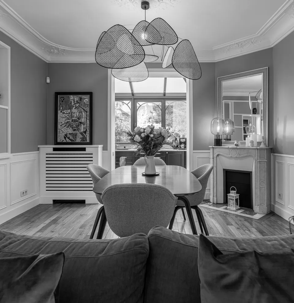 Studio Madame : architecte d’intérieur, Paris et Montmorency
Projet réalisé à Enghien-les-Bains.
Photographie d’intérieur (noir et blanc) : salle à manger avec vue sur la cuisine, nichée sous une verrière art déco. La salle à manger est composée d’une table ovale en noyer et de chaises en tissu, sous un lustre en cannage. Les murs et le plafond sont de type haussmannien, ornés de soubassements et moulures.