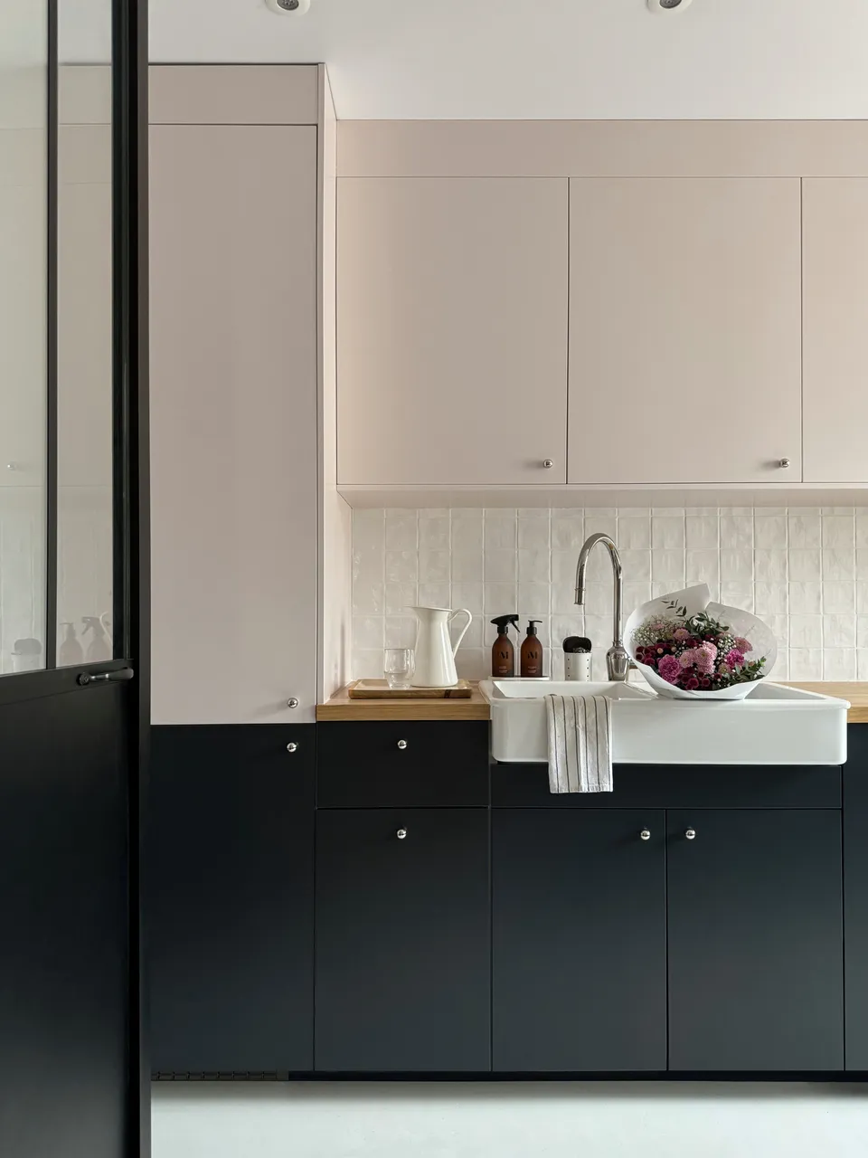 Studio Madame : architecte d’intérieur, Paris et Montmorency
Projet publié par ELLE DÉCORATION.
Projet réalisé à Enghien-les-Bains.
Photographie d’intérieur : cuisine habillée de façades rose pâle et bleu nuit et composée d’un plan de travail en chêne, d’un évier en céramique avec mitigeur chromé, d’une faïence blanche artisanale pour la crédence, de poignées chromées en forme de bulle et d’un sol en béton ciré gris clair. Des bouteilles de produit ménager, un pot en porcelaine, un plateau avec une carafe ancienne blanche et un verre, ainsi qu’un bouquet de fleurs dans des tons de rose sont posés sur le plan de travail et dans l’évier. Un torchon sèche sur le bord de l’évier. La porte permettant d’entrer dans la cuisine est une verrière industrielle noire.