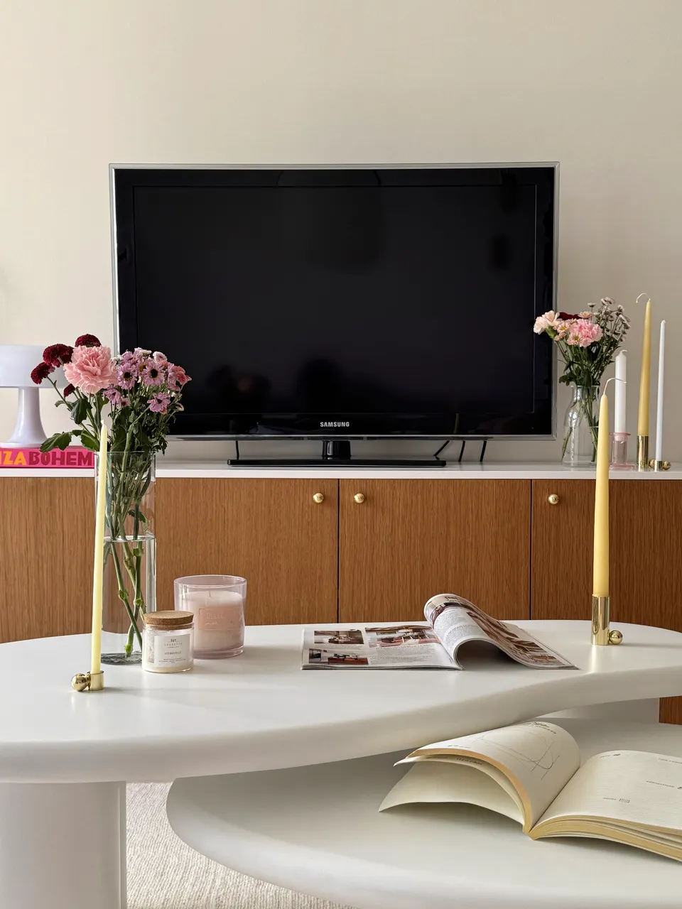 Studio Madame : architecte d’intérieur, Paris et Montmorency
Projet réalisé à Montmorency.
Photographie d’intérieur : salon composé d’un meuble TV rose et bois sur lequel reposent des bougeoirs, plusieurs bougies et un bouquet de fleurs. Un fauteuil blanc Roly Poly se tient juste à côté. Des tables basses asymétriques en ciment, sur lesquelles reposent un magazine, un carnet, plusieurs bougies et un bouquet de fleurs, sont placées devant.