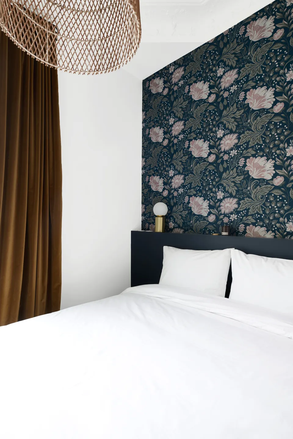 Studio Madame : architecte d’intérieur, Paris et Montmorency.
Projet réalisé à Boulogne-Billancourt. 
Photographie d’intérieur : chambre (vue sur coin nuit) peinte en blanc et bleu foncé (Ressource) et habillée d’un papier peint fleuri dans des tons de bleu et de rose.
Côté déco, le lit est recouvert d’une parure blanche, une lampe de chevet dorée est posée sur la tête de lit, et de grands rideaux ocres habillent les fenêtres.
Les autres murs sont peints en blanc (Ressource) et un parquet massif ancien est au sol.